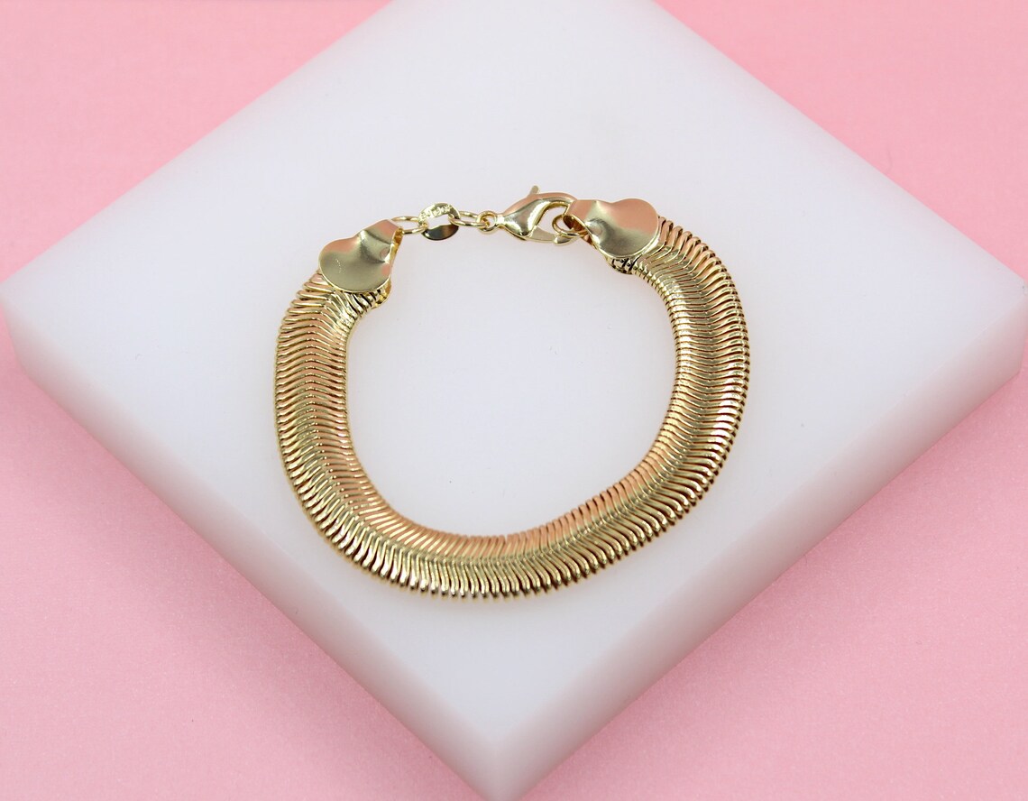 A thick gold herringbone snake chain bracelet rests on a white display block against a soft pink background.