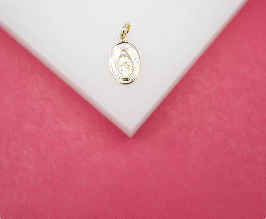 A small gold oval Miraculous Medal charm rests on a white display surface against a pink background.