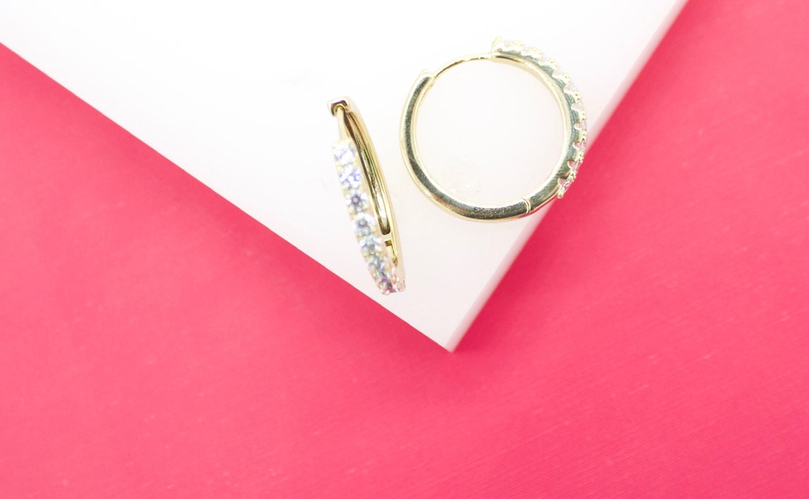 A pair of gold hoop earrings embellished with sparkling cubic zirconia stones rests on a white block against a bright pink background.