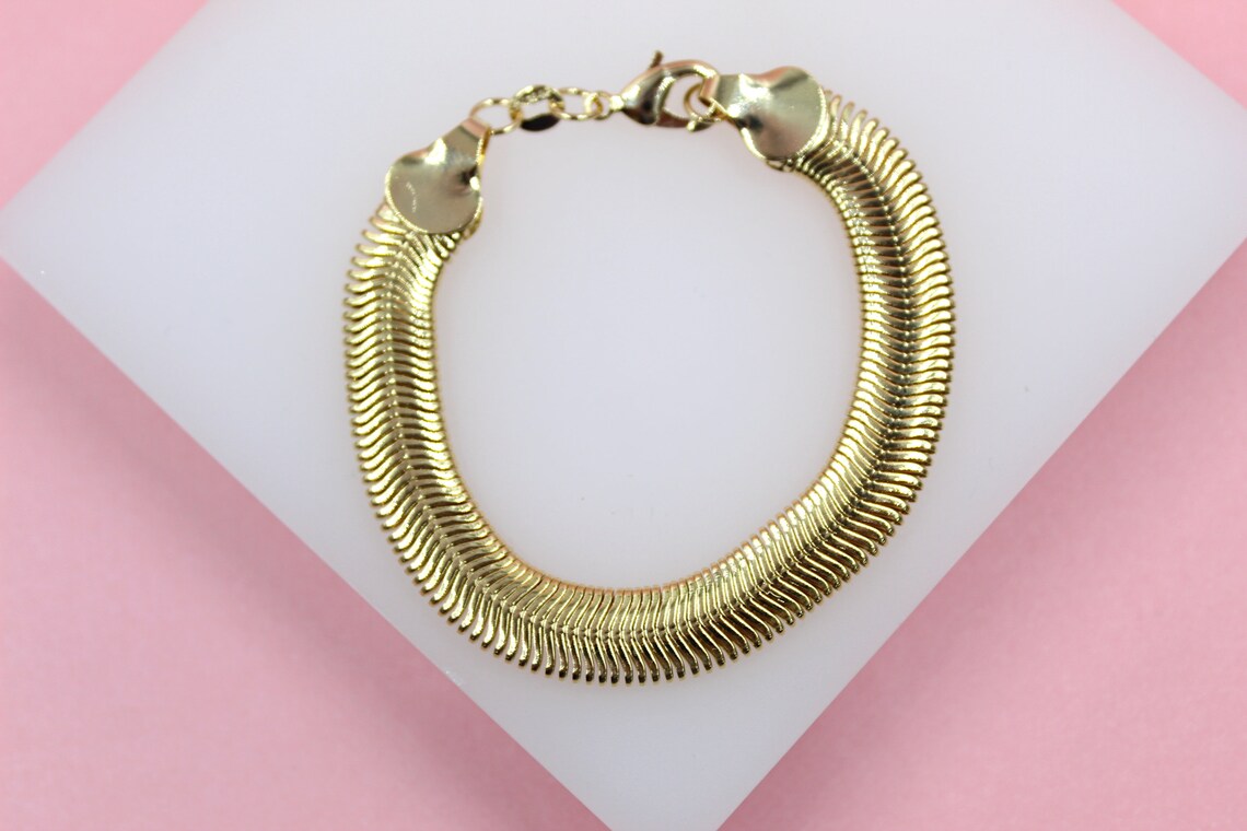 A thick gold snake chain bracelet rests on a white display stand against a soft pink background.