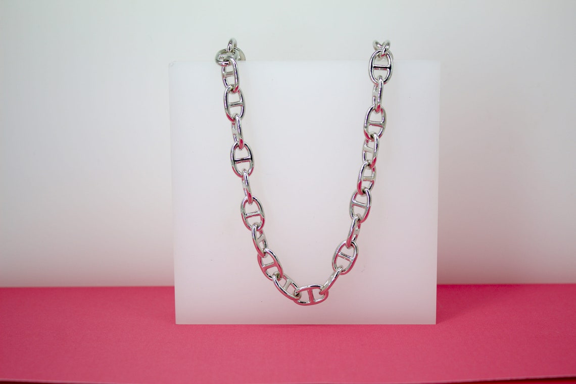 A thick silver anchor-link chain necklace rests draped over a white display block against a two-toned background.