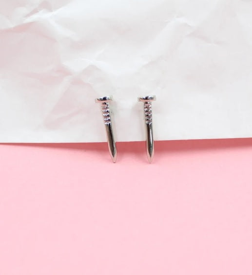 A pair of polished silver nail-shaped stud earrings rests against a textured white and soft pink background.