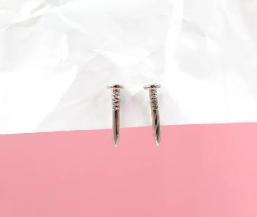 A pair of shiny silver nail-shaped stud earrings rest against a backdrop of white crumpled paper and a solid pink surface.