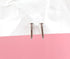A pair of shiny silver nail-shaped stud earrings rest against a backdrop of white crumpled paper and a solid pink surface.