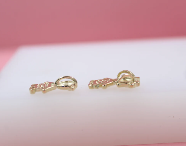 Two shiny gold teddy bear charms rest on a white surface against a pink background.