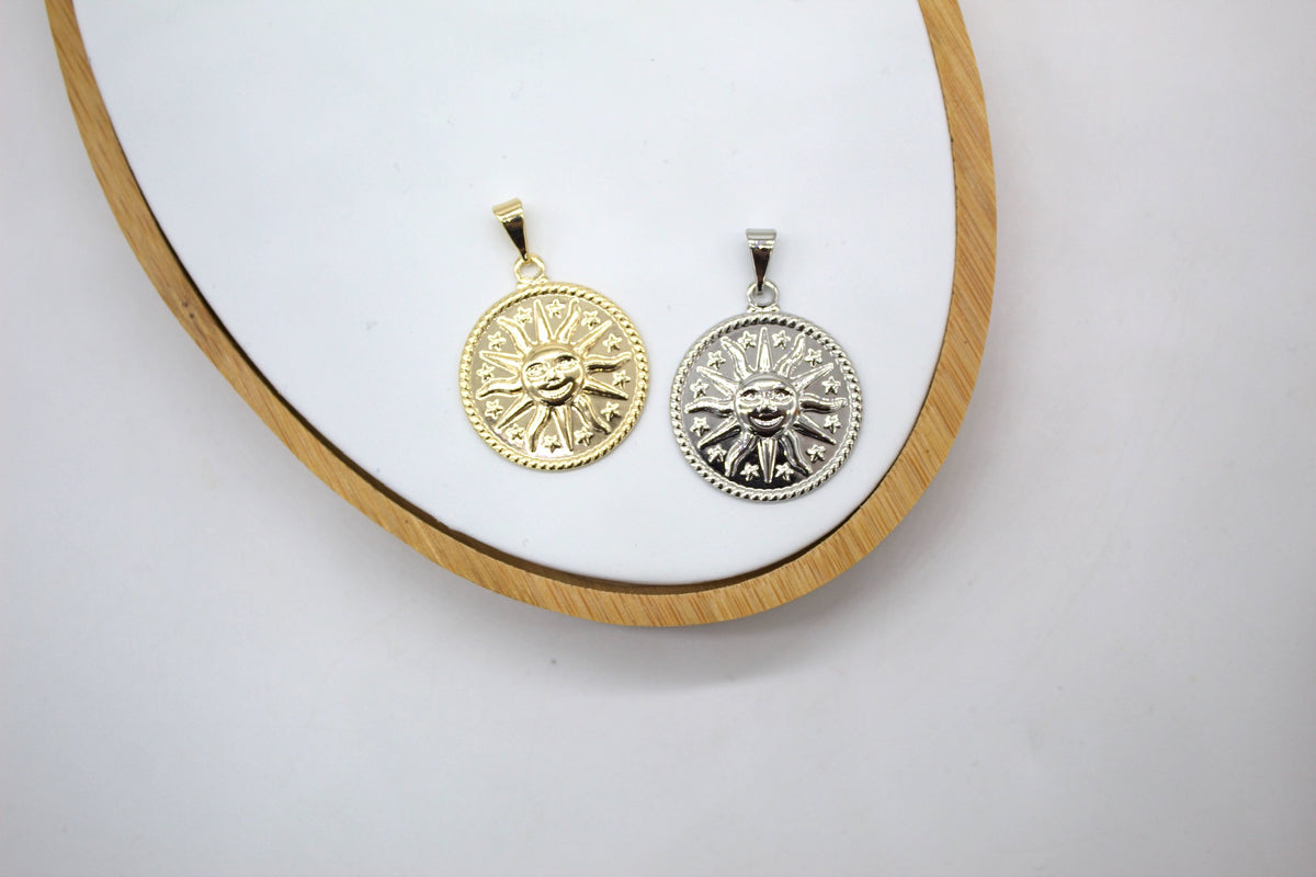 A gold and a silver celestial sun face pendant rest on a white display tray with a wooden rim.