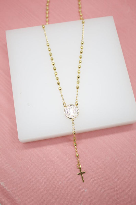 A delicate gold rosary necklace featuring beaded chains, a Saint Benedict medallion, and a crucifix rests on a white display block against a pink background.