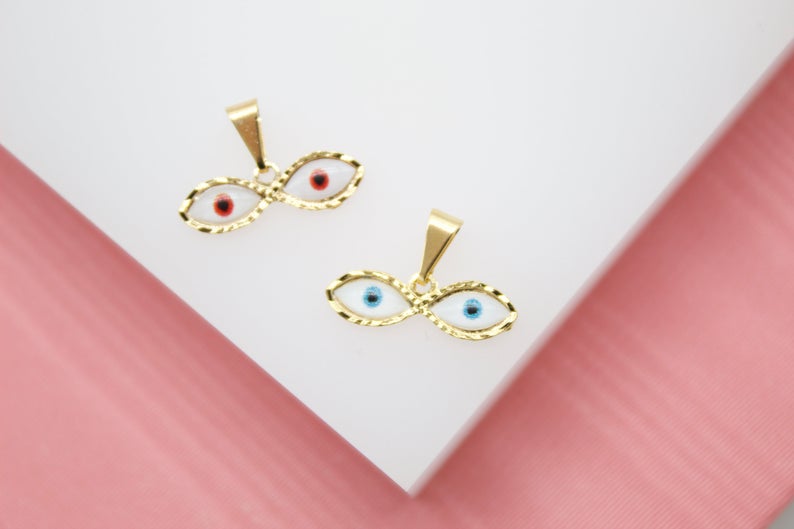 Two gold evil eye charms featuring white enamel and colorful pupils rest on a white surface against a pink background.