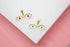 Two gold evil eye charms featuring white enamel and colorful pupils rest on a white surface against a pink background.