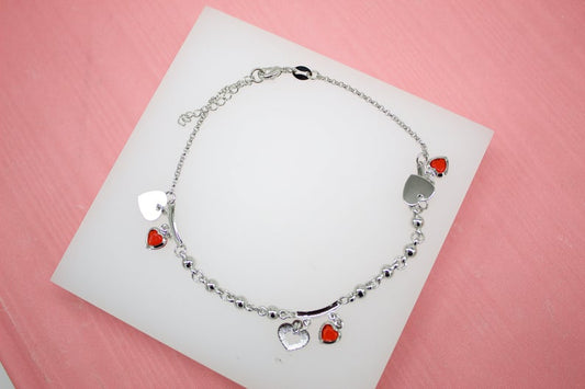 A delicate silver chain anklet featuring dangling red and silver heart charms rests on a white display surface.