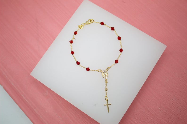 A delicate gold rosary bracelet features vibrant red crystal beads and a dangling cross pendant on a white display surface.