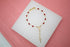 A delicate gold rosary bracelet features vibrant red crystal beads and a dangling cross pendant on a white display surface.