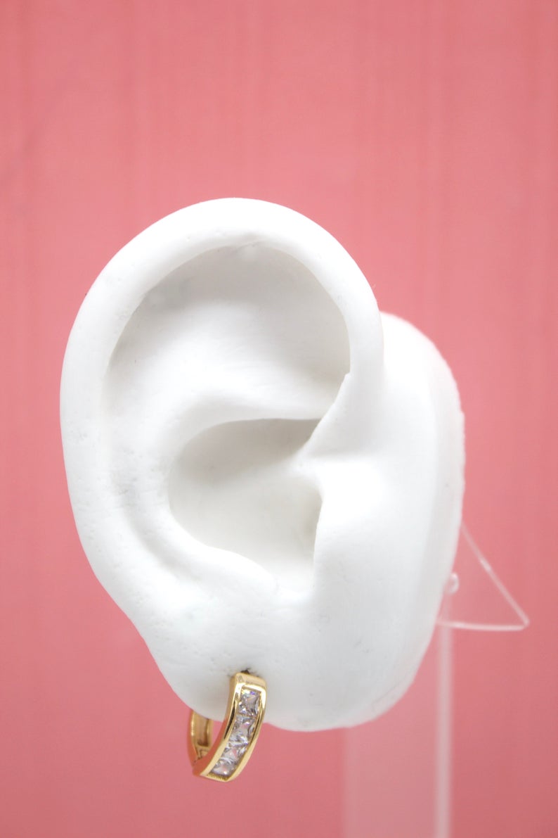 A white mannequin ear displays a sparkling gold huggie hoop earring adorned with small cubic zirconia stones against a solid pink background.
