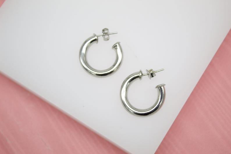 A pair of shiny silver chunky hoop earrings rests on a white display surface against a pink background.