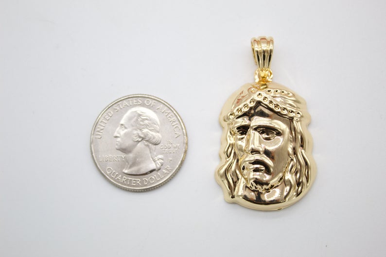 A shiny gold Jesus piece pendant rests next to a US quarter coin for scale on a light gray background.