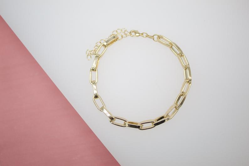 A chic gold paperclip chain bracelet rests against a pink and white surface.
