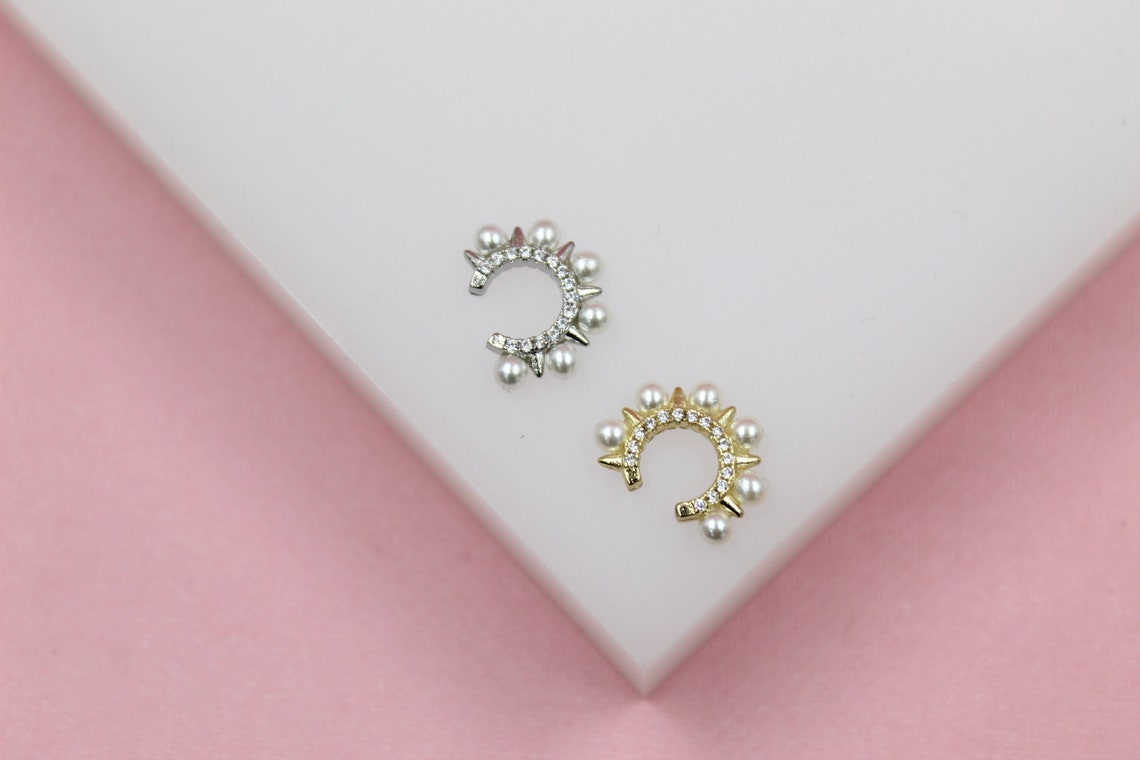 A silver and a gold ear cuff, both adorned with tiny pearls and sparkling crystals, rest on a white display against a pink background.