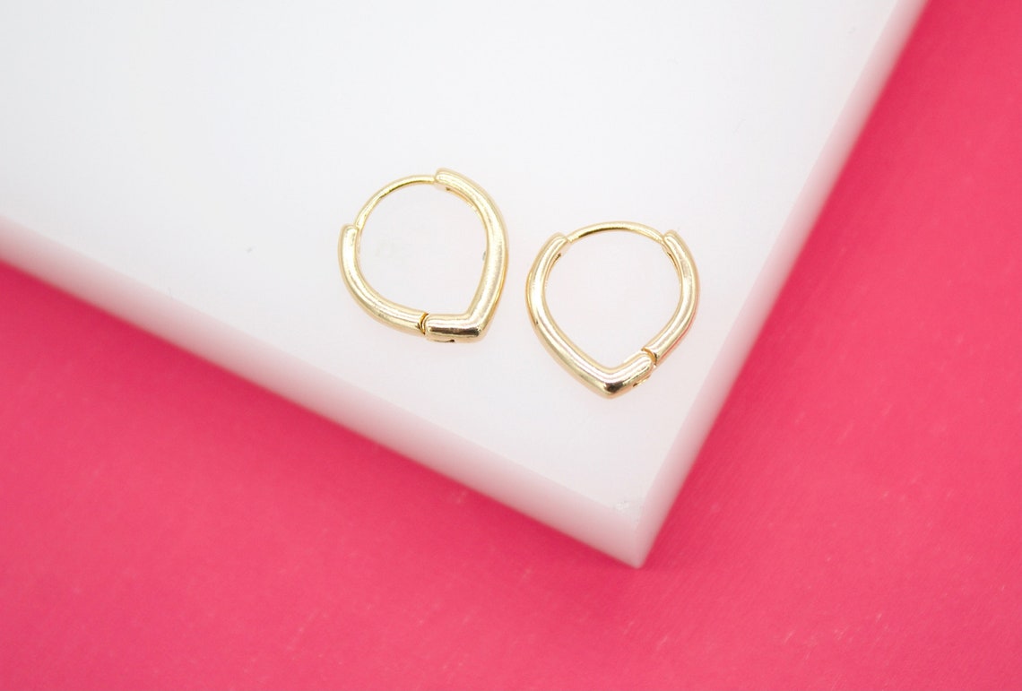 A pair of small gold hoop earrings with a subtle heart shape rests on a white block against a bright pink background.