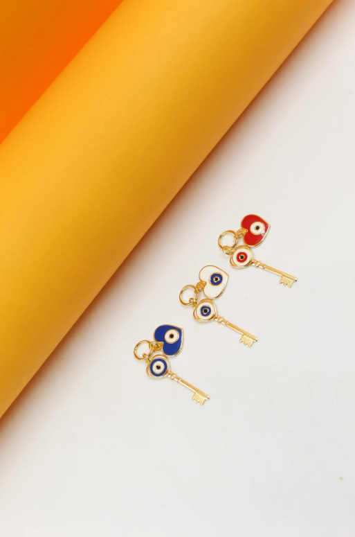 Three gold key charms featuring evil eye and heart pendants in blue, white, and red rest on a white surface next to a bright orange cylindrical roll.