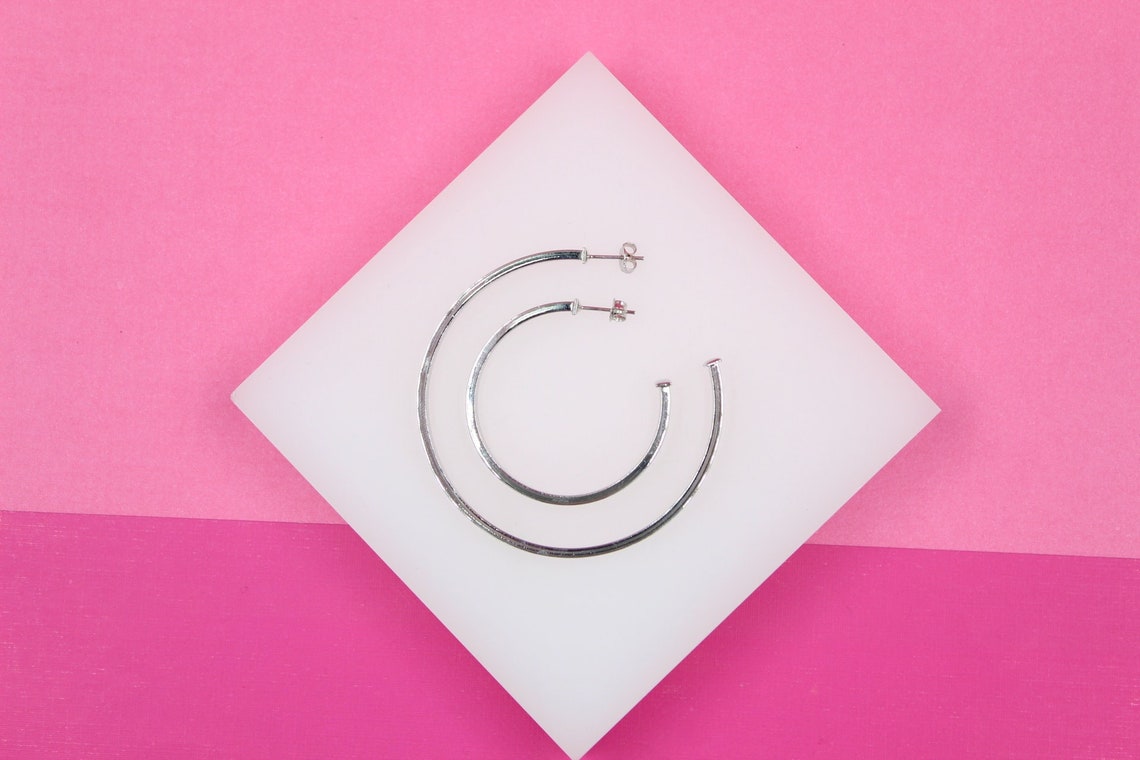 Two sterling silver hoop earrings rest on a white square display against a vibrant pink background.