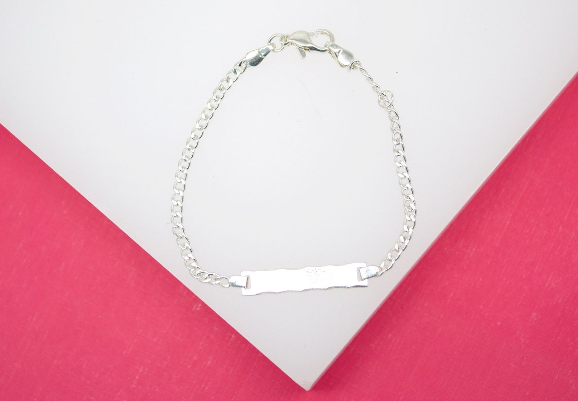 A delicate silver identity bracelet with a blank ID plate rests against a white and pink background.