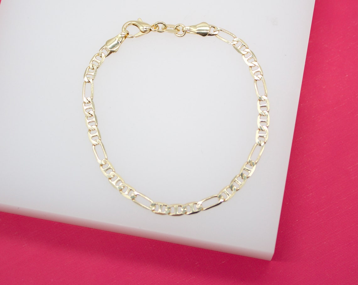 A delicate gold Figaro link chain bracelet rests on a white display surface against a vibrant pink background.