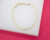 A delicate gold Figaro link chain bracelet rests on a white display surface against a vibrant pink background.