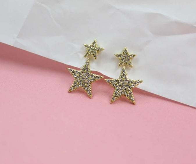 A pair of dazzling gold star-shaped earrings embedded with sparkling cubic zirconia rests on a pink and white surface.