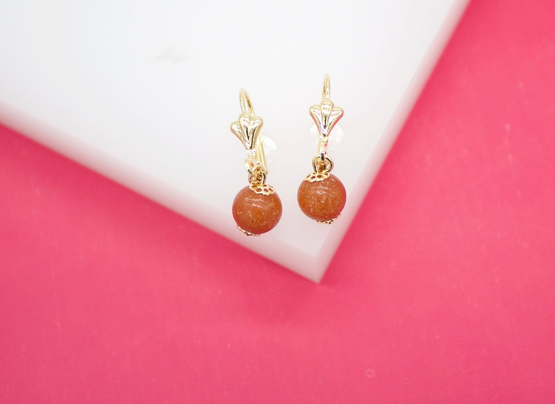 A pair of gold dangle earrings featuring sparkling orange gemstone beads rests against a white block on a vibrant pink background.