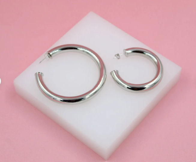Two polished silver hoop earrings rest on a white square display against a pink background.