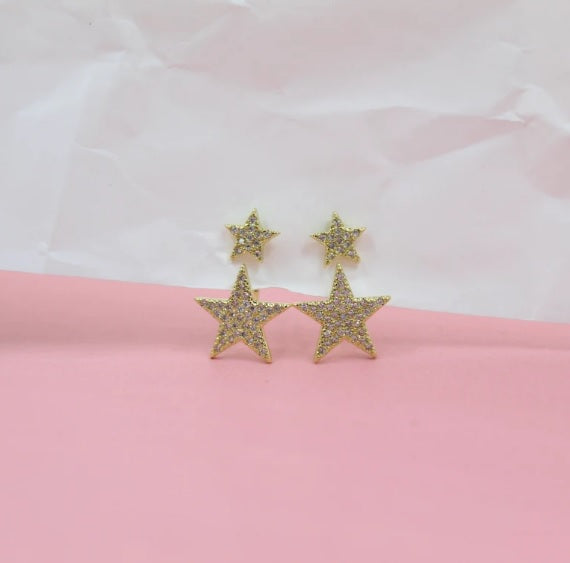 Sparkling gold star stud earrings rest against a soft pink and white textured backdrop.