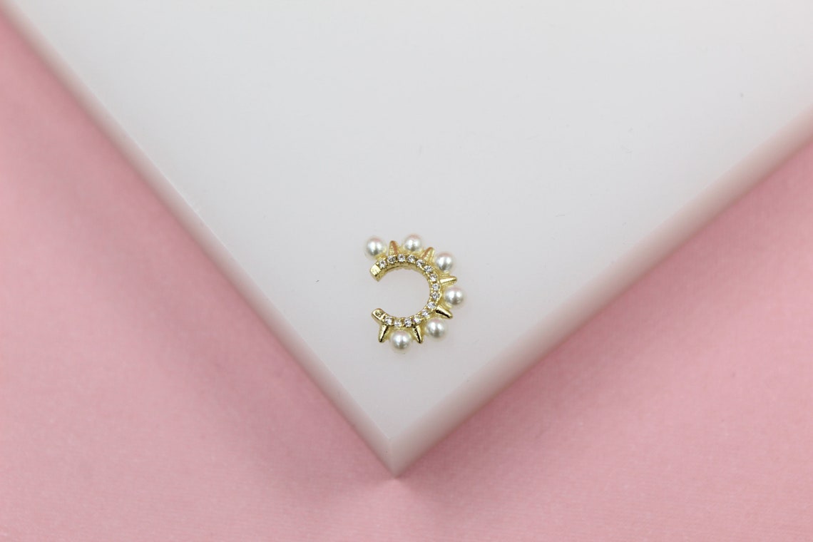 A gold cuff earring features sparkling pave crystals and delicate pearl accents against a white and pink background.