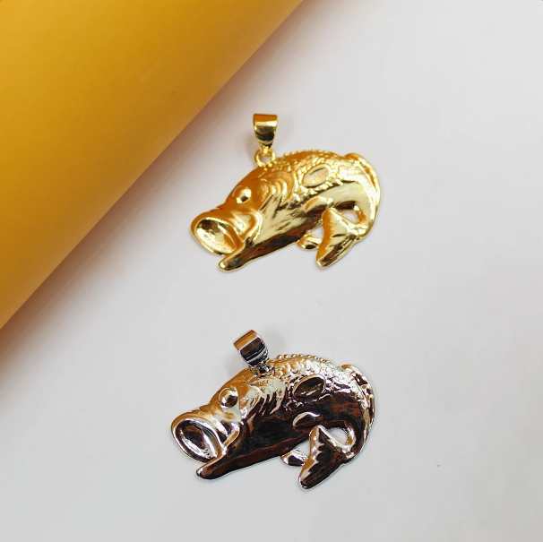 Two polished fish-shaped pendants, one gold and one silver, rest on a white surface next to a yellow cylinder.