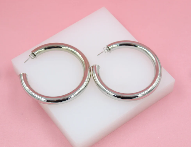 A pair of thick silver hoop earrings rests on a white display block against a soft pink background.