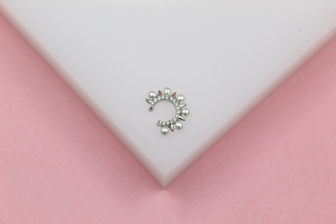A detailed silver ear cuff features sparkling cubic zirconia stones, small spikes, and delicate pearls, resting on a white display against a soft pink background.