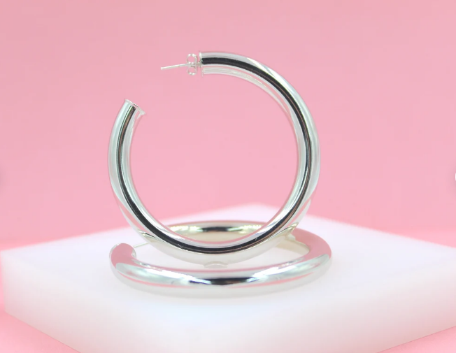 A pair of thick silver hoop earrings rests on a white display block against a soft pink background.