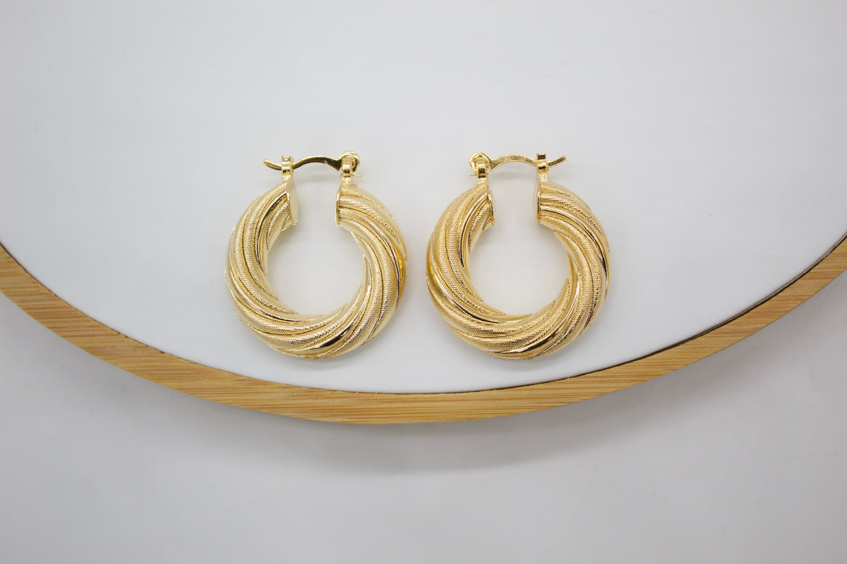 A pair of textured gold twist hoop earrings rests on a white display surface with a wooden rim.
