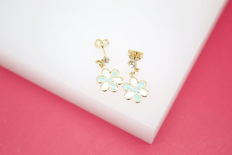 A pair of gold drop earrings featuring light blue and white enamel flower charms rests on a white and pink surface.