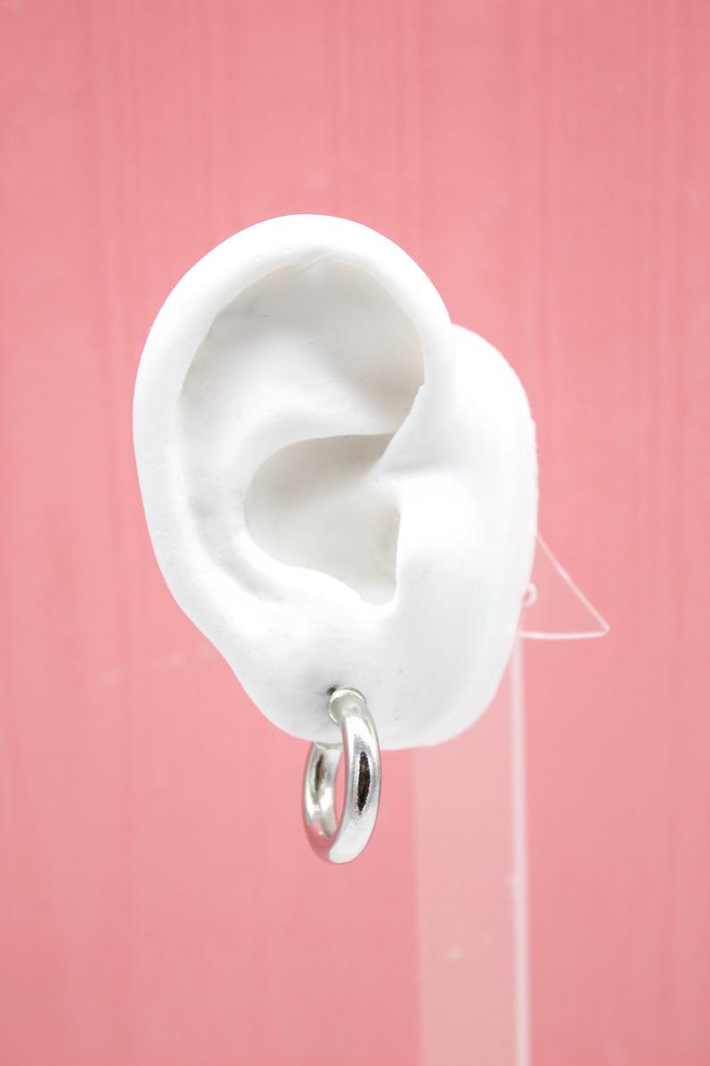 A white mannequin ear displays a polished silver hoop earring against a textured pink background.