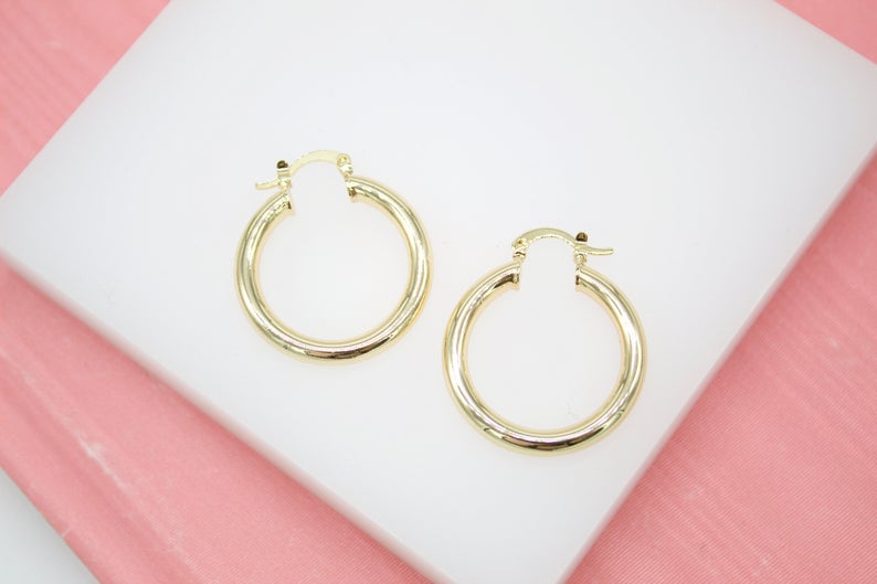 A pair of polished gold hoop earrings rests neatly on a white display surface against a soft pink background.