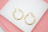 A pair of polished gold hoop earrings rests neatly on a white display surface against a soft pink background.