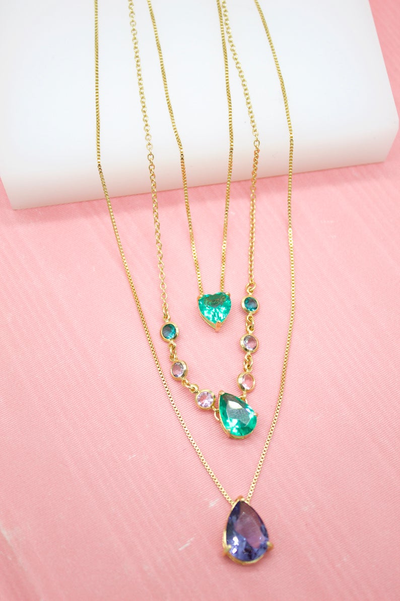 Three layered gold necklaces featuring green heart and teardrop gemstone pendants rest on a pink surface.