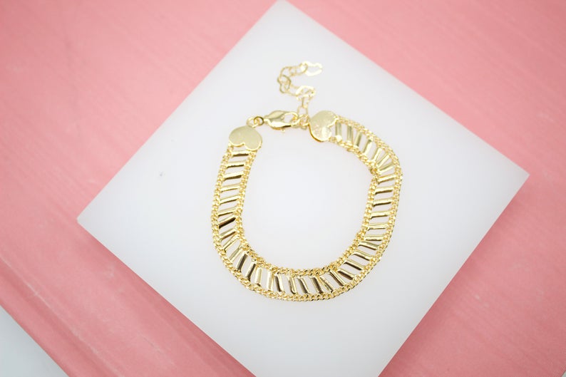 A gold-plated link bracelet rests on a white display surface against a pink background.