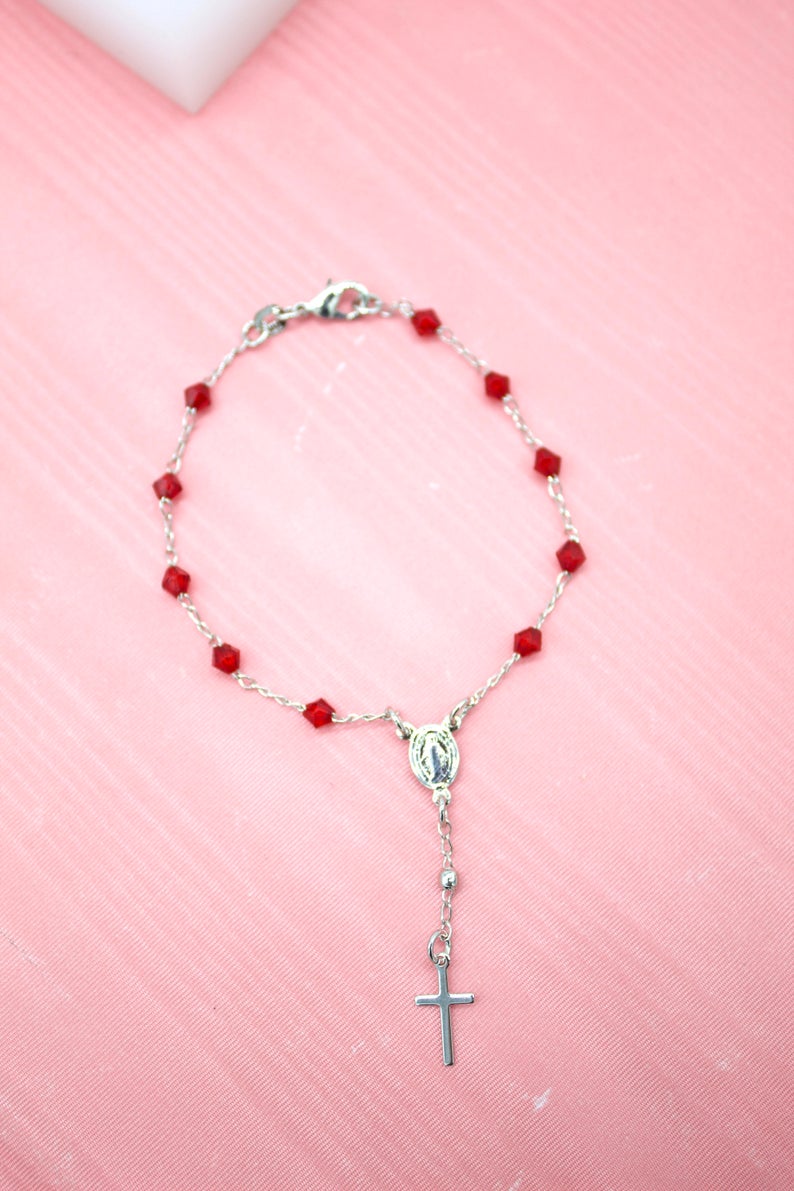 A silver rosary bracelet featuring faceted red crystal beads lies flat on a light pink surface.