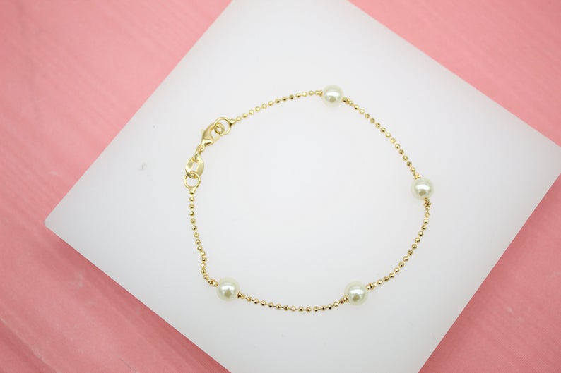 A delicate gold beaded bracelet with scattered white pearls rests on a white display surface against a pink background.