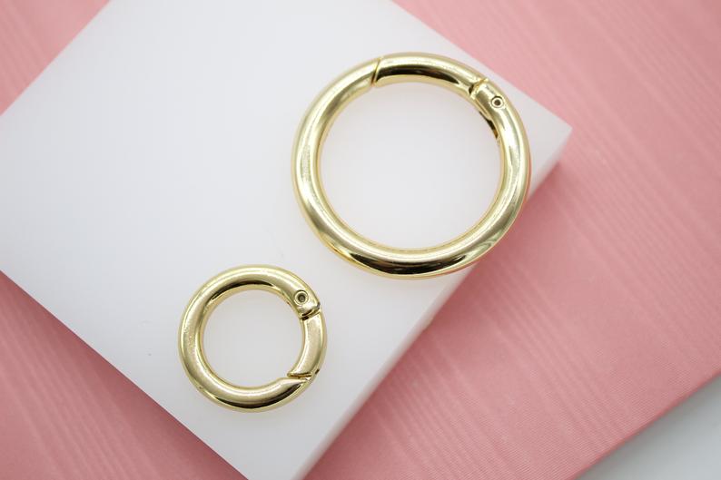 Two polished gold spring ring clasps rest on a white display block against a pink background.