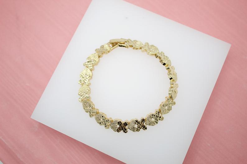 A gold hug and kiss XO bracelet rests on a white display stand against a pink backdrop.
