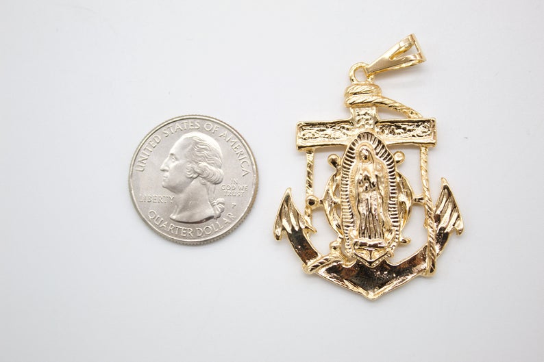 A gold anchor pendant featuring Our Lady of Guadalupe rests beside a US quarter coin to show its scale.