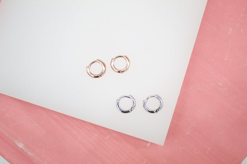 A pair of rose gold and a pair of silver hoop earrings rest on a white surface against a pink background.