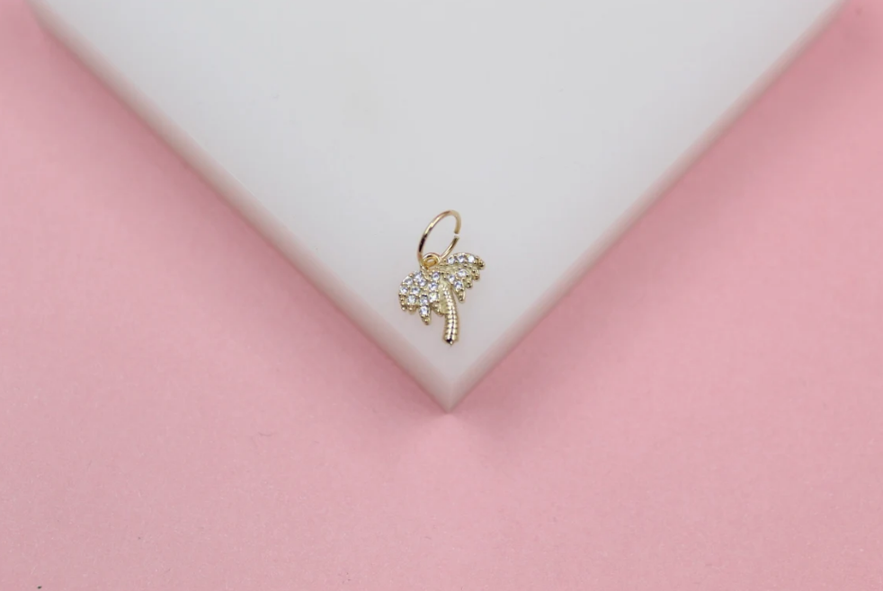 A gold palm tree charm embellished with sparkling cubic zirconia stones rests delicately on a white display surface against a soft pink background.